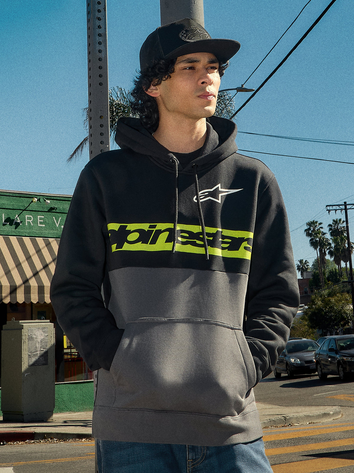 Felpa con cappuccio Alpinestars Parallel, streetwear lifestyle, design a blocchi di colore nero e gunmetal, striscia verde lime sul petto con logo Alpinestars, logo Astars bianco, cappuccio con cordino, indossata da un uomo in un contesto urbano all'aperto.