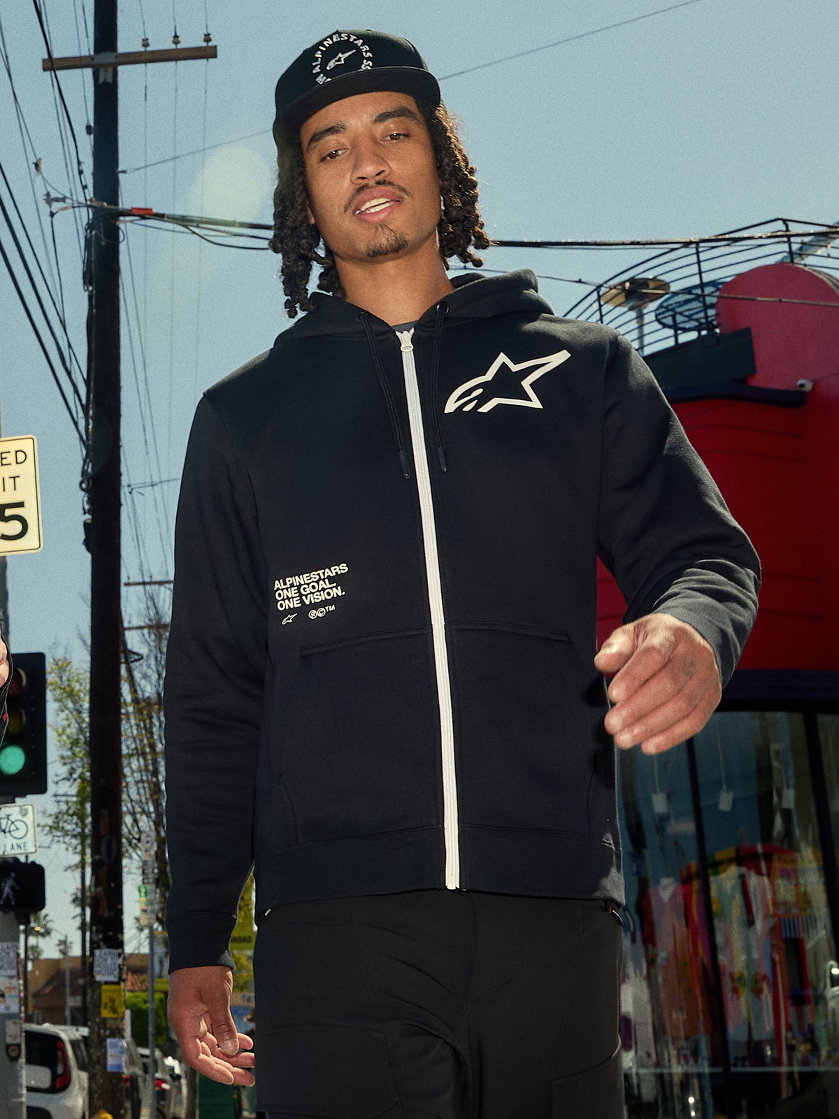 Felpa con cappuccio Alpinestars Transverse, felpa in pile nera con zip frontale, zip bianca e logo Astars bianco sul petto, stampa con testo 'One Goal One Vision', stile streetwear casual, indossata da un uomo in un ambiente urbano all'aperto