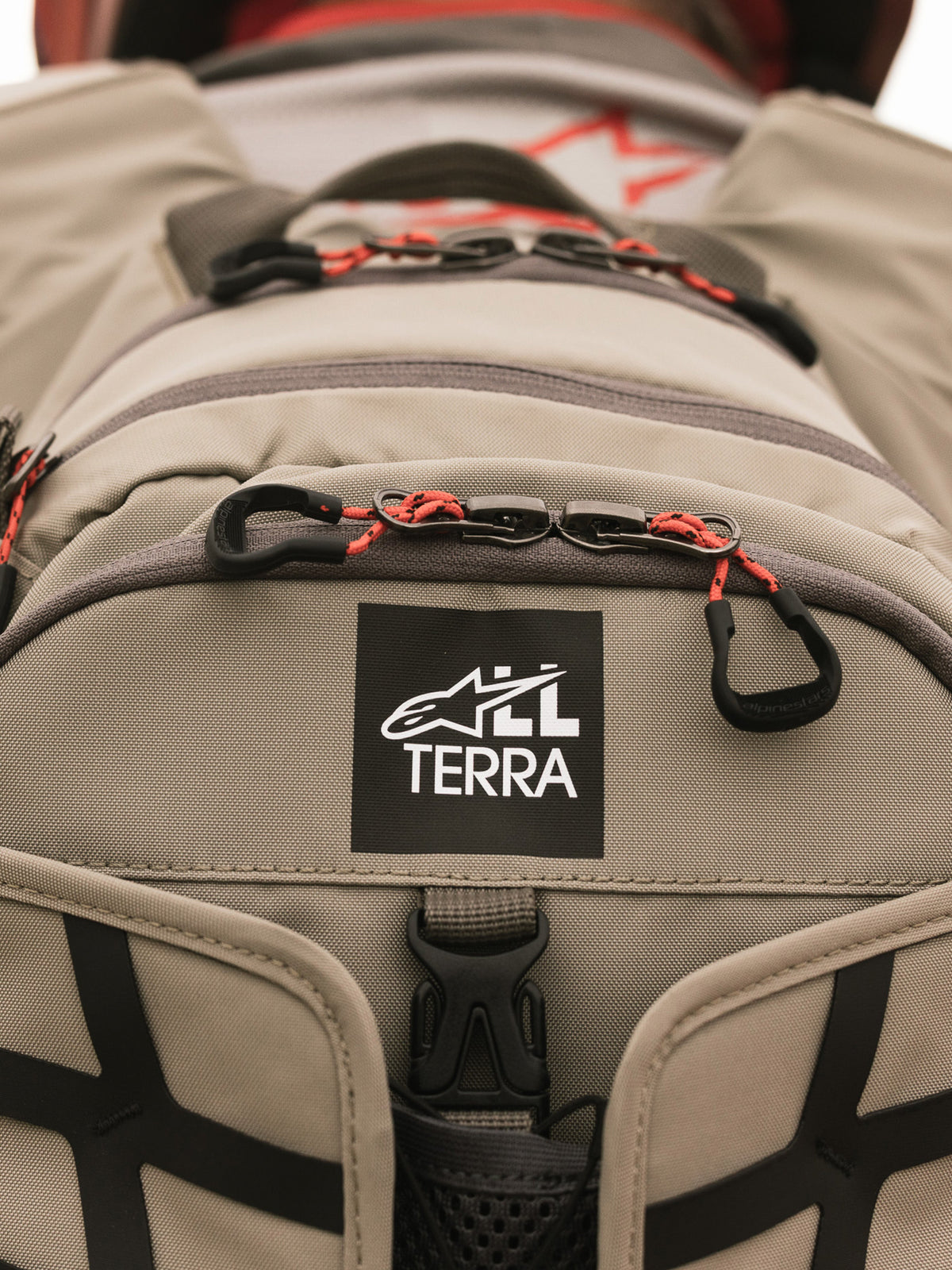 Alpinestars AT-15 Back Pack, Aluminum Walnut, vista ravvicinata della toppa con logo All Terra su tessuto tecnico color sabbia, con tiretti delle cerniere in cordino arancione e dettagli di costruzione robusti