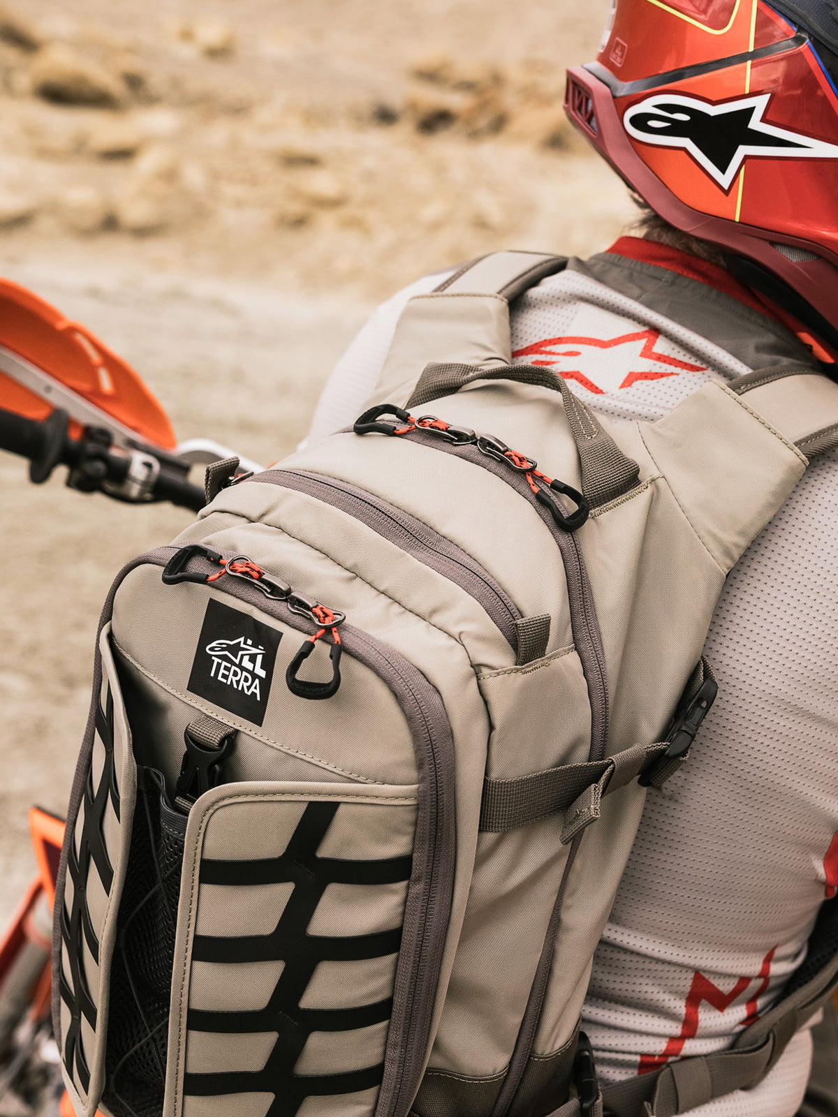 Zaino Alpinestars AT-15, zaino da moto Adventure, Aluminum Walnut e nero, indossato da un pilota in un contesto desertico, con tiralampo rossi, patch con logo TERRA e fettuccia esterna nera, abbinato a un casco rosso e maglia Alpinestars bianca