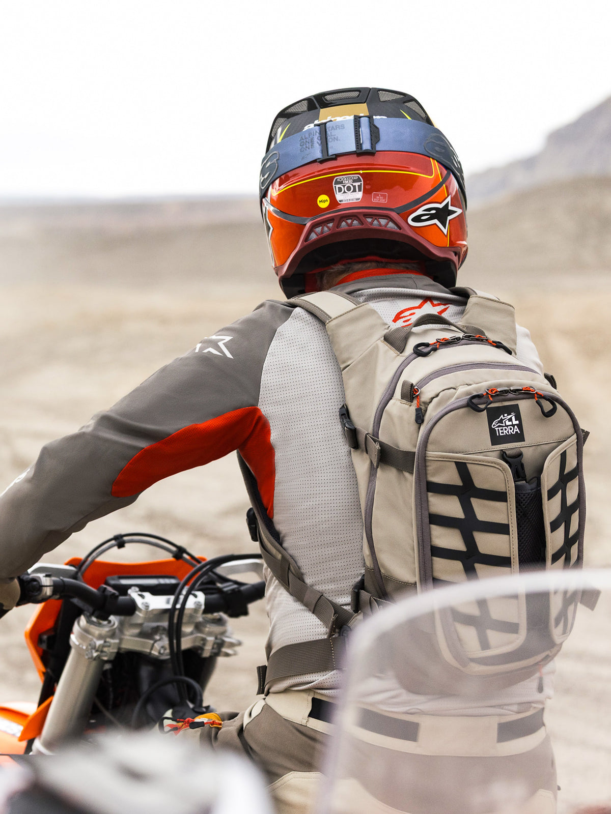 Zaino Alpinestars AT-15 in colore Aluminum Walnut, indossato da un motociclista adventure in un paesaggio desertico. Presenta una robusta struttura in tessuto beige con cinghie nere, dettagli delle cerniere arancioni e marchio 'All Terra'. Abbinato a un casco rosso e arancione e abbigliamento grigio per touring fuoristrada.