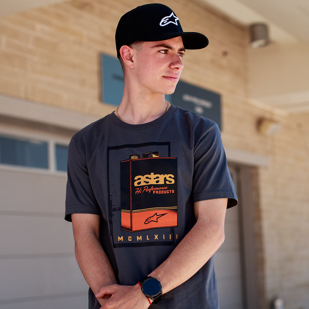Alpinestars Galun CSF Tee, t-shirt a maniche corte, grigio, grafica frontale con lattina di gas vintage, marchio 'astars' e numeri romani 'MCMLXIII', indossata da un uomo con cappellino nero con logo, stile casual motorsport