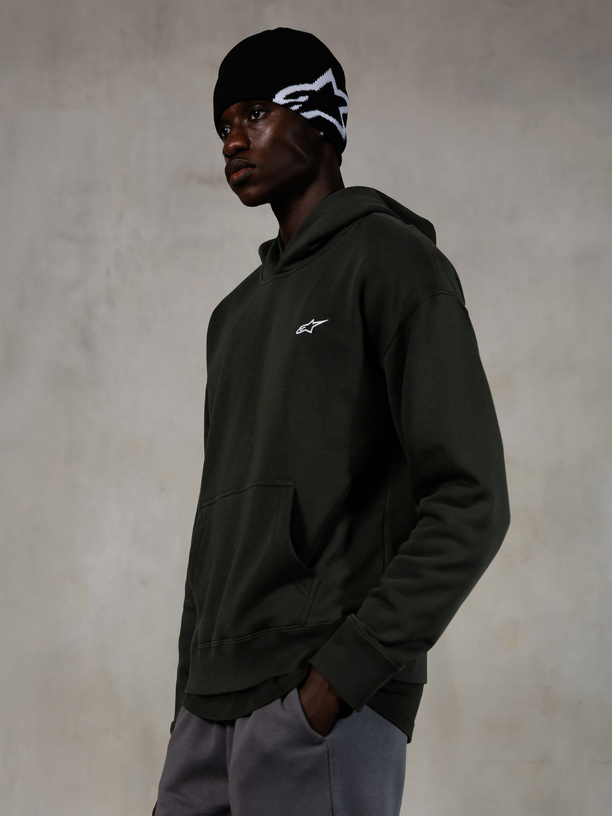 Felpa con cappuccio Alpinestars Essentials Ageless, Off Black, modello che indossa streetwear casual, pile grigio antracite con finitura stone wash e piccolo logo a stella bianco, abbinata a un berretto Alpinestars nero e pantaloni grigi