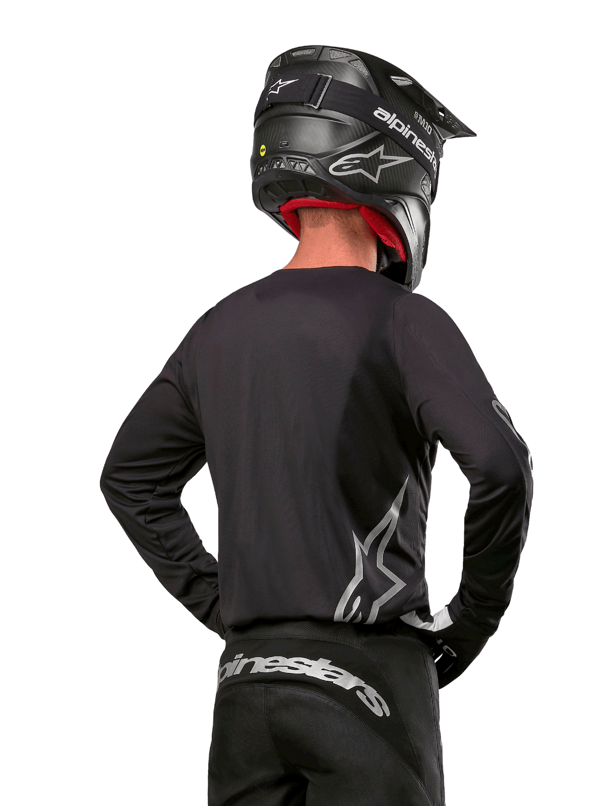 Pilota di motocross visto di spalle con maglia Alpinestars Fluid Graphite 2024 in nero e argento, caratterizzata da un grande logo argento sul lato, casco nero con trama in carbonio e pantaloni neri, abbigliamento professionale per corse fuoristrada