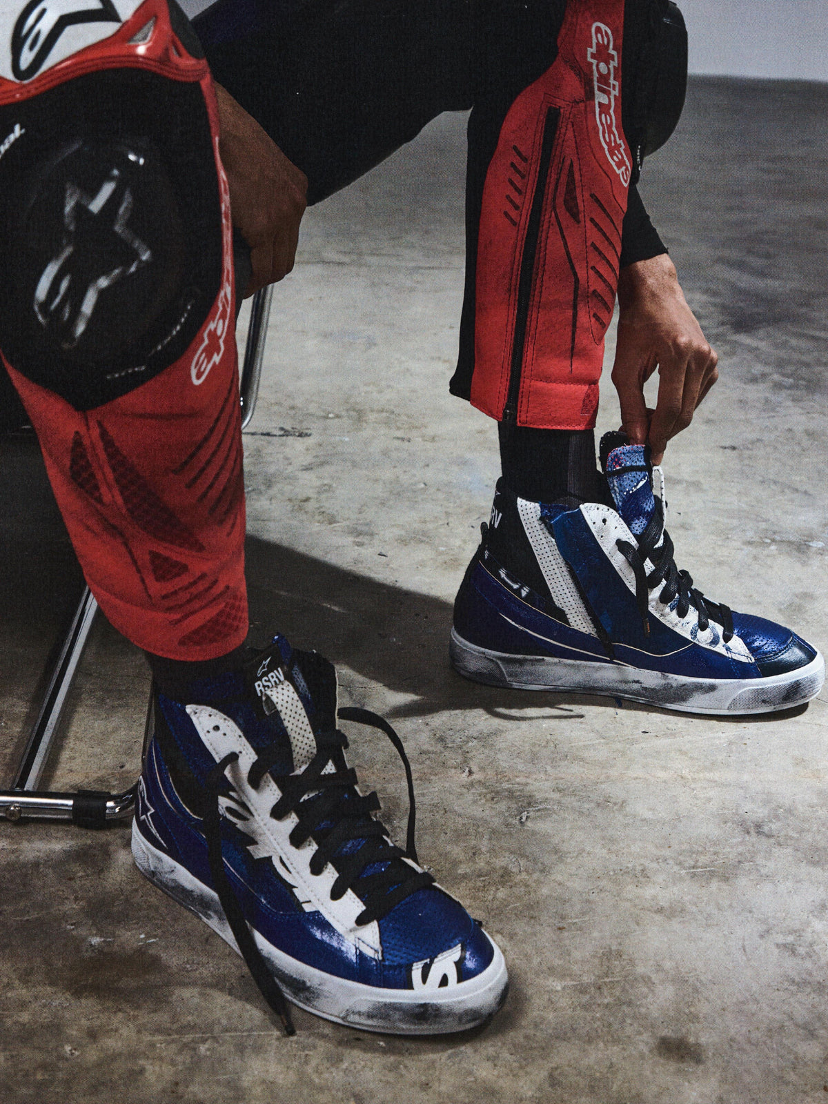 Alpinestars RSRV Legacy Shoe, scarpe alte blu e bianche con lacci neri, indossate con pantaloni protettivi rossi e neri, suola consumata, texture lucida, su pavimento in cemento grigio