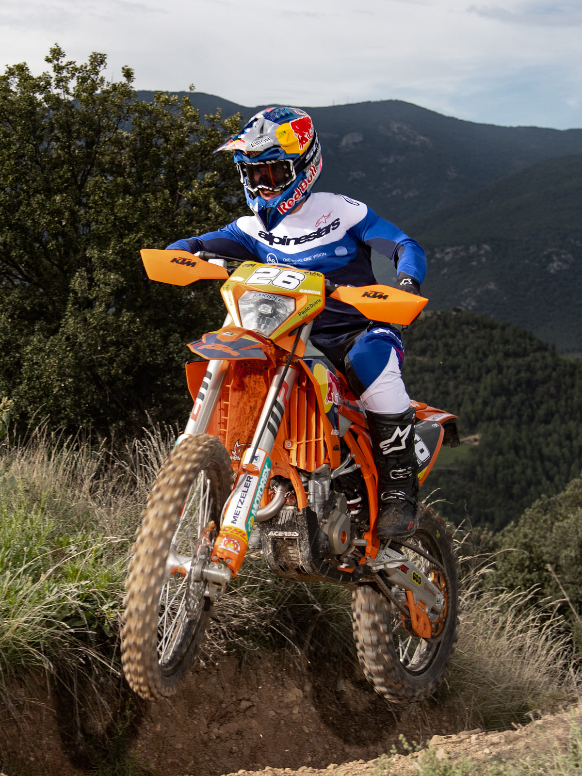 Pilota di enduro su una moto arancione, maglia Alpinestars Pro-Dura in Navy Bright Blue White, pantaloni neri e bianchi e stivali Alpinestars neri, mentre percorre un sentiero accidentato con montagne e alberi sullo sfondo.
