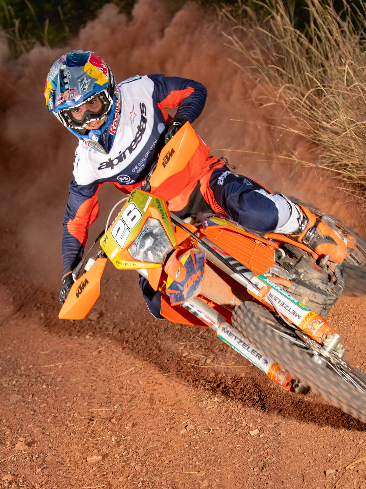 Pilota di enduro in azione su una moto da cross arancione, indossa la maglia Alpinestars Pro-Dura nei colori blu navy, arancione acceso e bianco, caratterizzata da un design leggero e traspirante con branding audace per prestazioni fuoristrada.