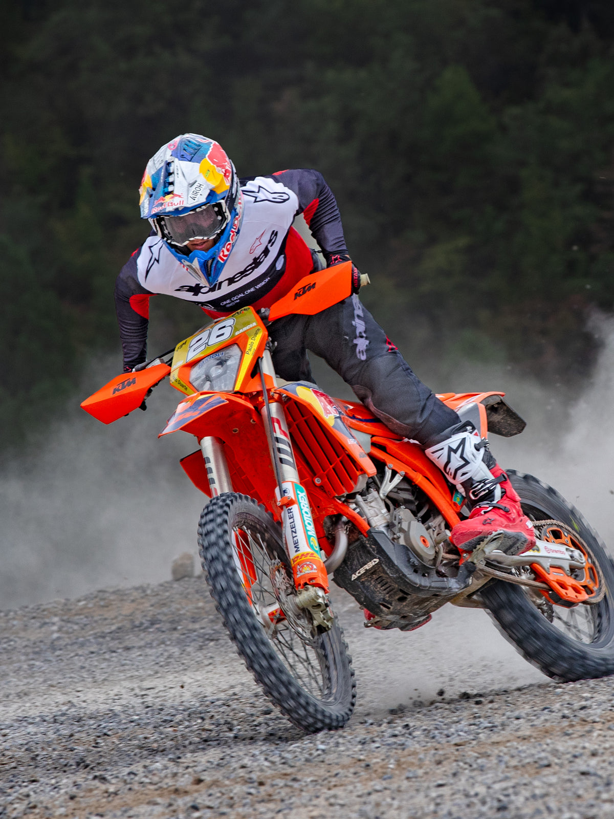 Pilota di motocross in azione su una moto KTM arancione, indossa l'abbigliamento Alpinestars Pro-Dura con maglia rossa, bianca e nera, pantaloni grigio scuro, stivali rossi e bianchi e casco da corsa Red Bull, eseguendo una curva stretta su un sentiero sterrato