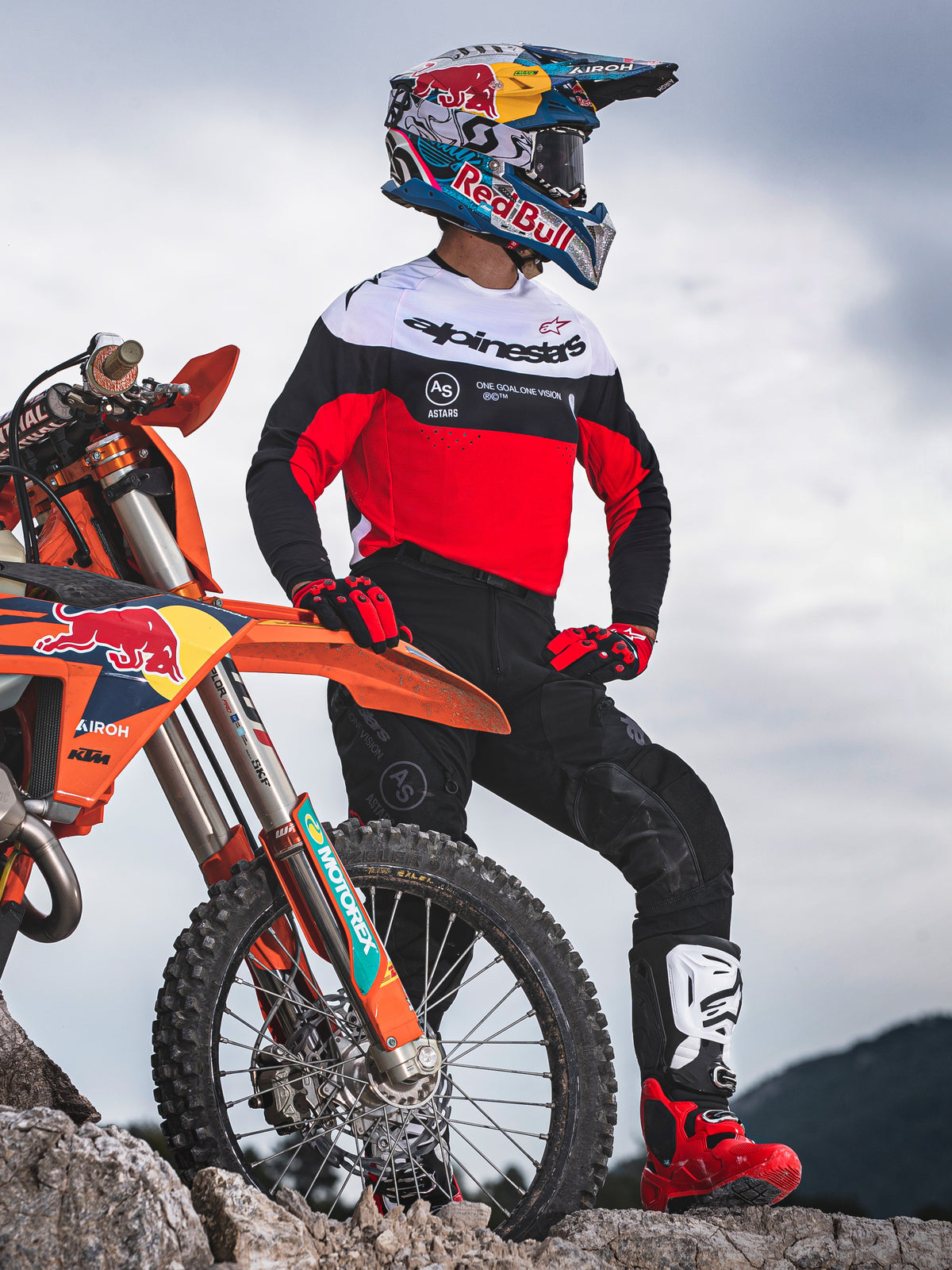 Pilota di motocross accanto a una moto off-road arancione su terreno roccioso, indossa abbigliamento Alpinestars Pro-Dura con maglia rossa, bianca e nera e pantaloni neri, guanti rossi e neri, stivali bianchi e rossi e casco Red Bull, completo professionale da enduro