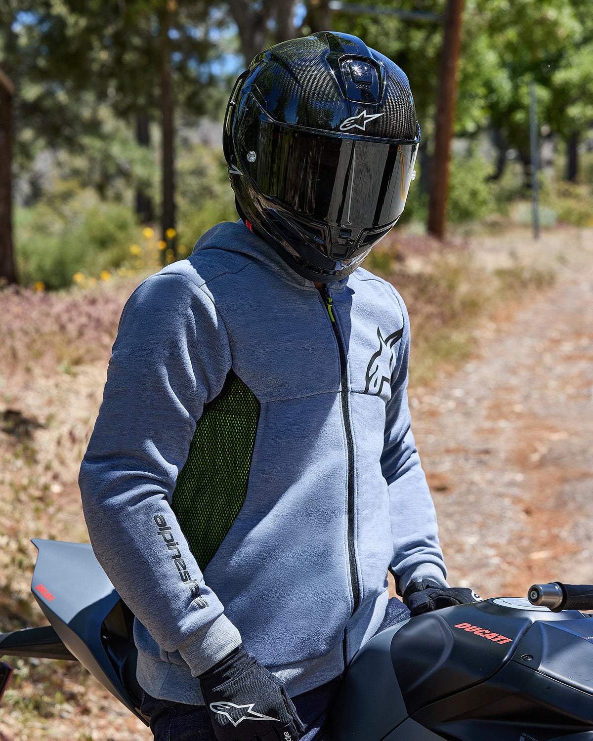 Motociclista con felpa Alpinestars Chrome Air Sport Hoodie in grigio mélange, pannelli laterali in mesh verde lime e loghi neri, abbinata a casco in fibra di carbonio e guanti neri, seduto su una moto Ducati in un ambiente all'aperto.