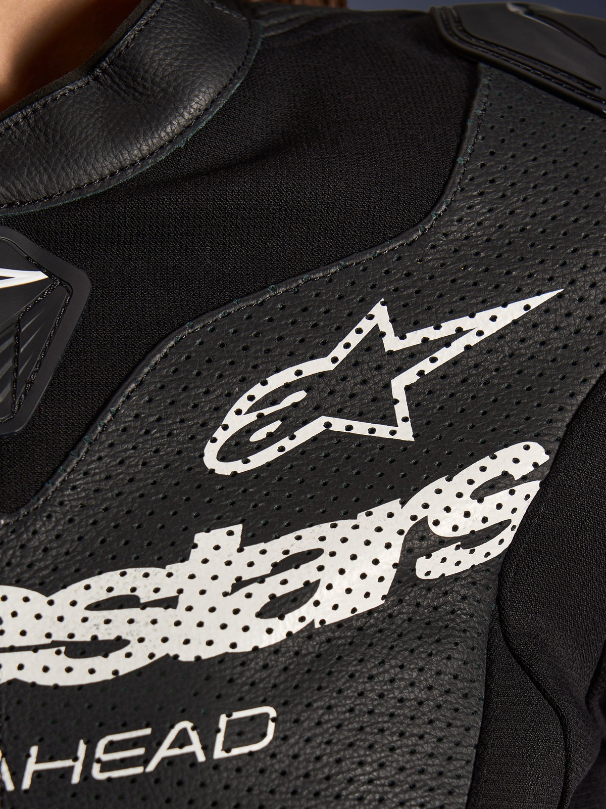 Primo piano della tuta in pelle Alpinestars Stella GP-R7 2 Pezzi in nero, caratterizzata da pannelli in pelle traforata, un grande logo Alpinestars bianco sul petto e sezioni in tessuto elastico traspirante.