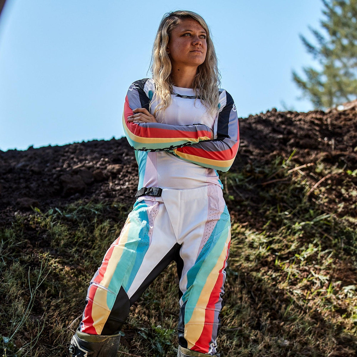 Pilota di motocross donna con pantaloni Alpinestars Stella Techstar 2024 e maglia coordinata, design Black Multicolor con base bianca e strisce blu, gialle e rosse, in piedi con le braccia incrociate su pista sterrata, abbigliamento professionale da gara fuoristrada