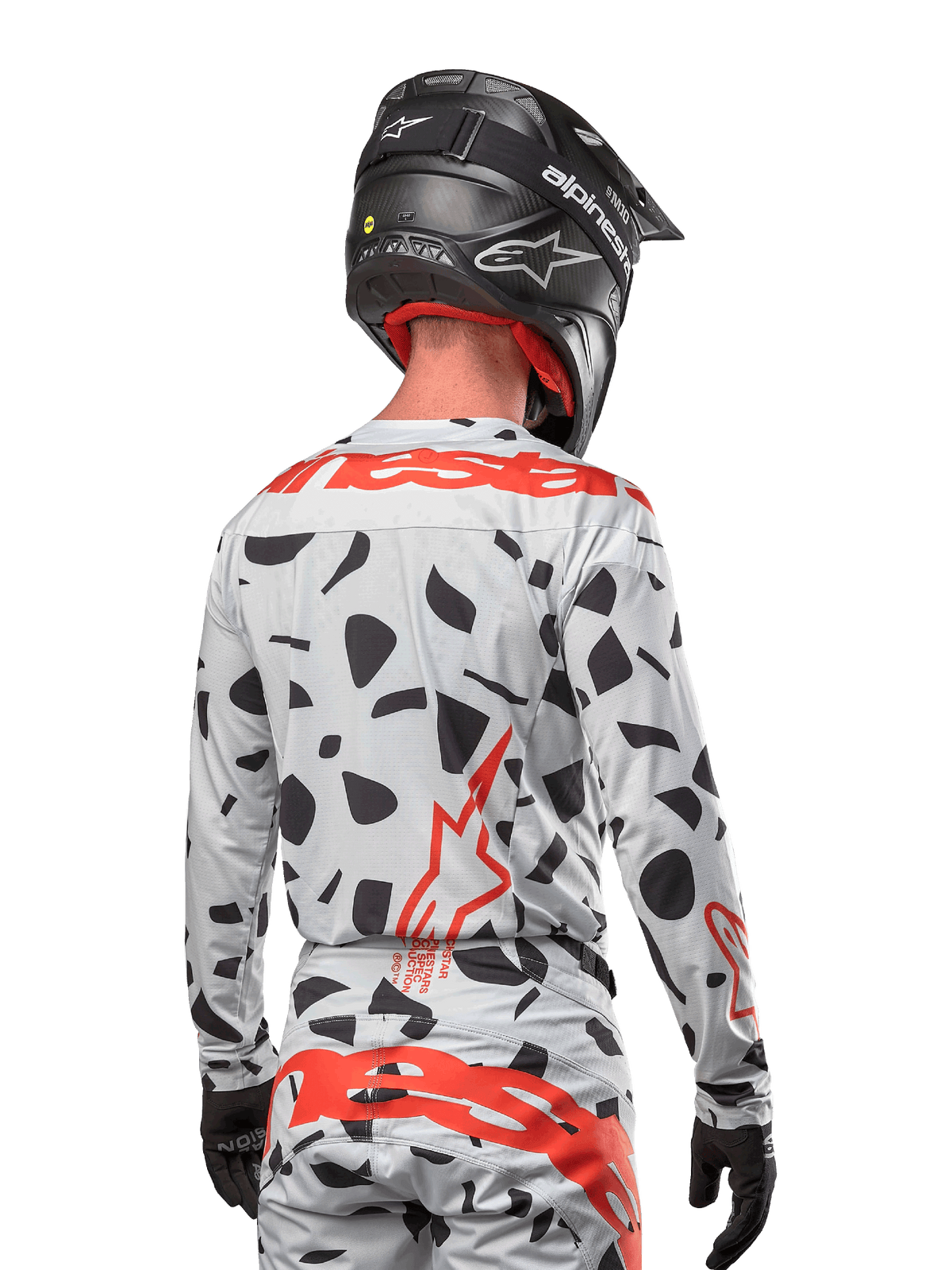Pilota di motocross visto di spalle che indossa la maglia Alpinestars 2024 Techstar Rantera in Haze Gray Camo, con base grigio chiaro e macchie grafiche nere, vivaci loghi Alpinestars rossi su schiena e spalle, abbinata a pantaloni coordinati e casco nero.