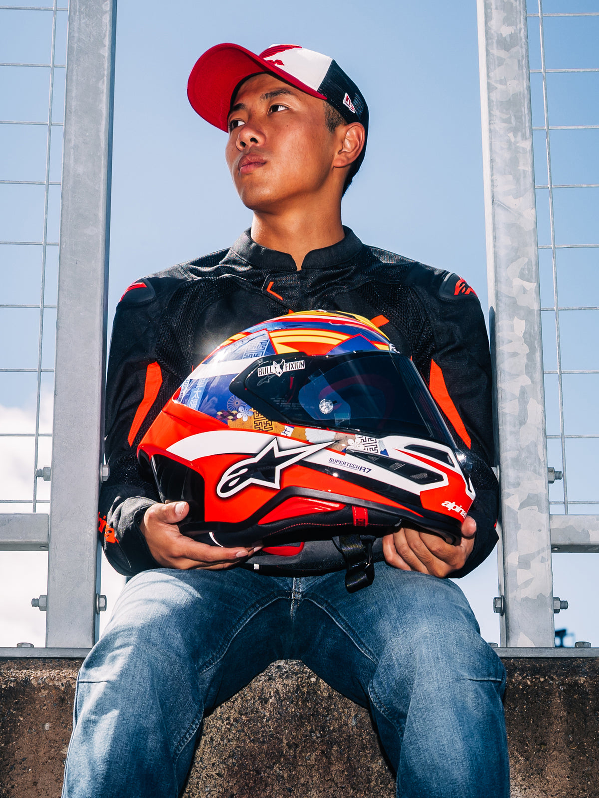 Alpinestars Casco S-R7 Taiyo Furusato, casco integrale da moto racing, grafica rossa, bianca e blu, tenuto dal pilota Taiyo Furusato con giacca nera, vista all'aperto a bordo pista