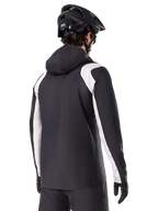 Alpinestars Giacca Antipioggia A-Dura, giacca antipioggia da mountain bike, nera con pannelli bianchi sulle maniche, vista posteriore del ciclista con casco da MTB nero e guanti, costruzione tecnica impermeabile a 2,5 strati, abbigliamento da mountain bike