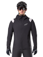 Alpinestars A-Dura Rain Jacket, giacca antipioggia MTB, nero con pannelli bianchi sulle spalle, indossata con casco MTB nero, occhiali da sole e guanti, design impermeabile con cappuccio e cerniera frontale, abbigliamento sportivo outdoor per il ciclismo