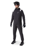 Alpinestars A-Dura Rain Jacket, giacca antipioggia da mountain bike nera, con accenti bianchi sulle spalle, indossata da un ciclista con casco da MTB, occhiali da sole, guanti e pantaloni neri coordinati, abbigliamento tecnico da ciclismo per ogni condizione meteo