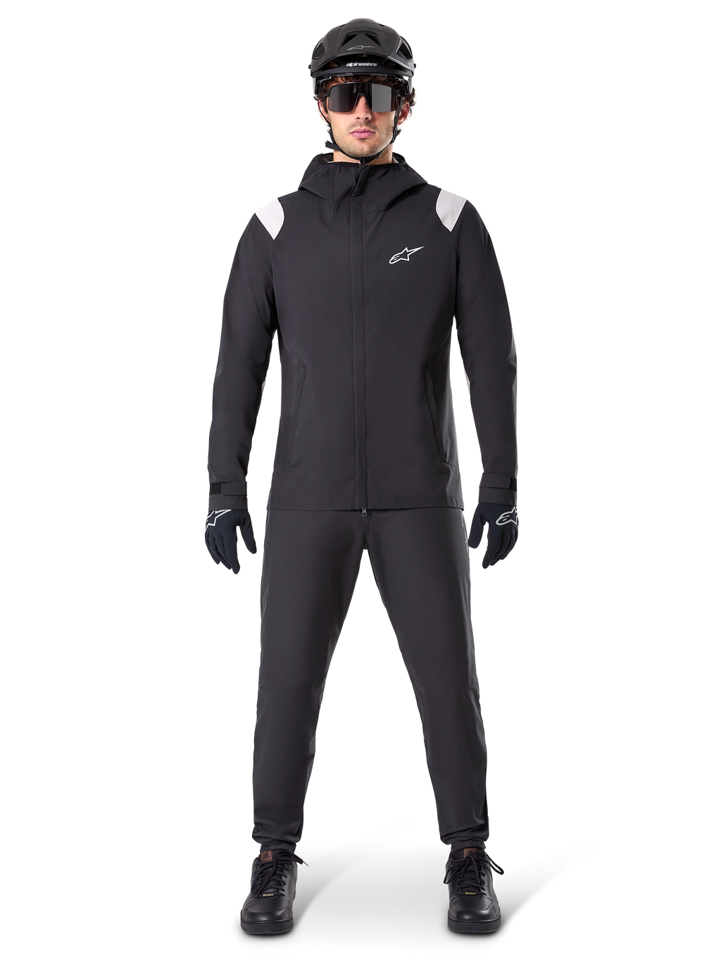 Giacca Antipioggia Alpinestars A-Dura, giacca antipioggia da mountain bike, nera con accenti bianchi sulle spalle e logo bianco sul petto, indossata con pantaloni neri, casco nero, occhiali da sole e guanti, abbigliamento coordinato da mountain bike per il maltempo
