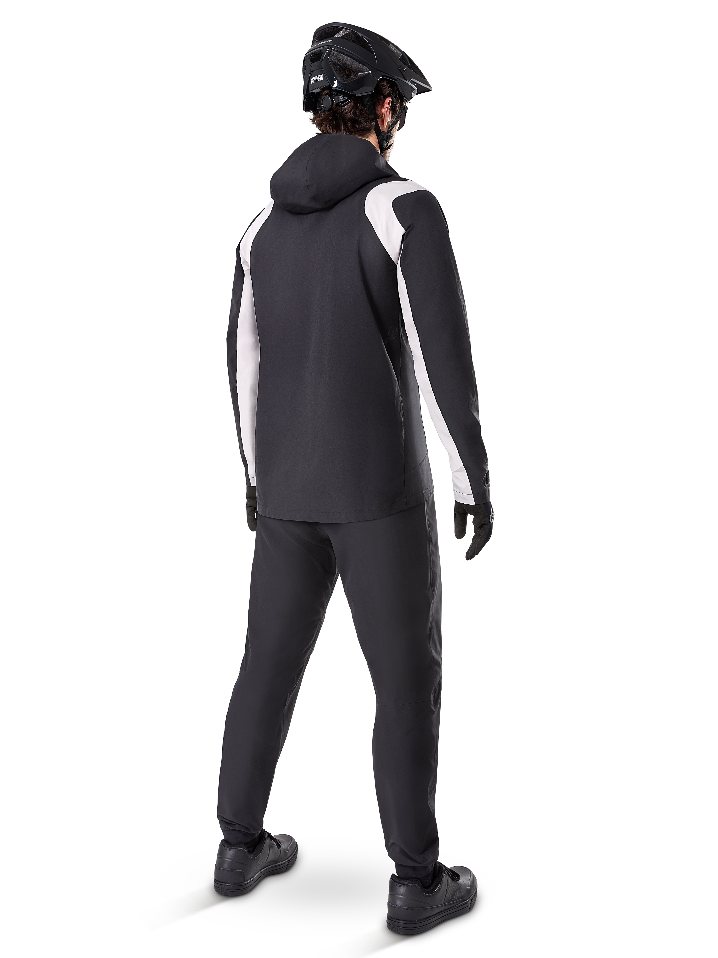 Alpinestars Giacca Antipioggia A-Dura, vista posteriore di un ciclista che indossa la giacca da MTB impermeabile bianca e nera, pantaloni da ciclismo neri coordinati, casco e guanti neri, equipaggiamento tecnico per mountain bike in condizioni di pioggia