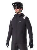 Alpinestars A-Dura Rain Jacket, giacca antipioggia da mountain bike, nera con inserti grigio chiaro sulle spalle, indossata da un ciclista con casco da MTB nero e occhiali da sole, logo bianco sul petto, costruzione impermeabile, coordinata con pantaloni e guanti neri per ciclismo fuoristrada