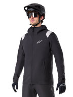 Alpinestars A-Dura Rain Jacket, giacca antipioggia da mountain bike, nera con inserti grigio chiaro sulle spalle, indossata da un ciclista con casco da MTB nero e occhiali da sole, logo bianco sul petto, costruzione impermeabile, coordinata con pantaloni e guanti neri per ciclismo fuoristrada