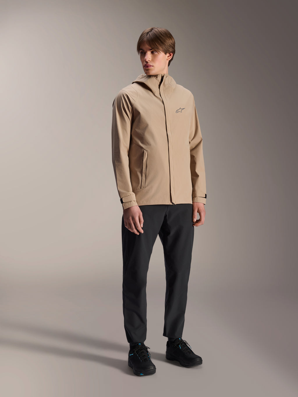 Modello uomo che indossa la giacca antipioggia Alpinestars A-Dura in Light Brown, abbinata a pantaloni e scarpe neri, giacca antipioggia minimalista per mountain bike outdoor con design con cappuccio e un piccolo logo sul petto.