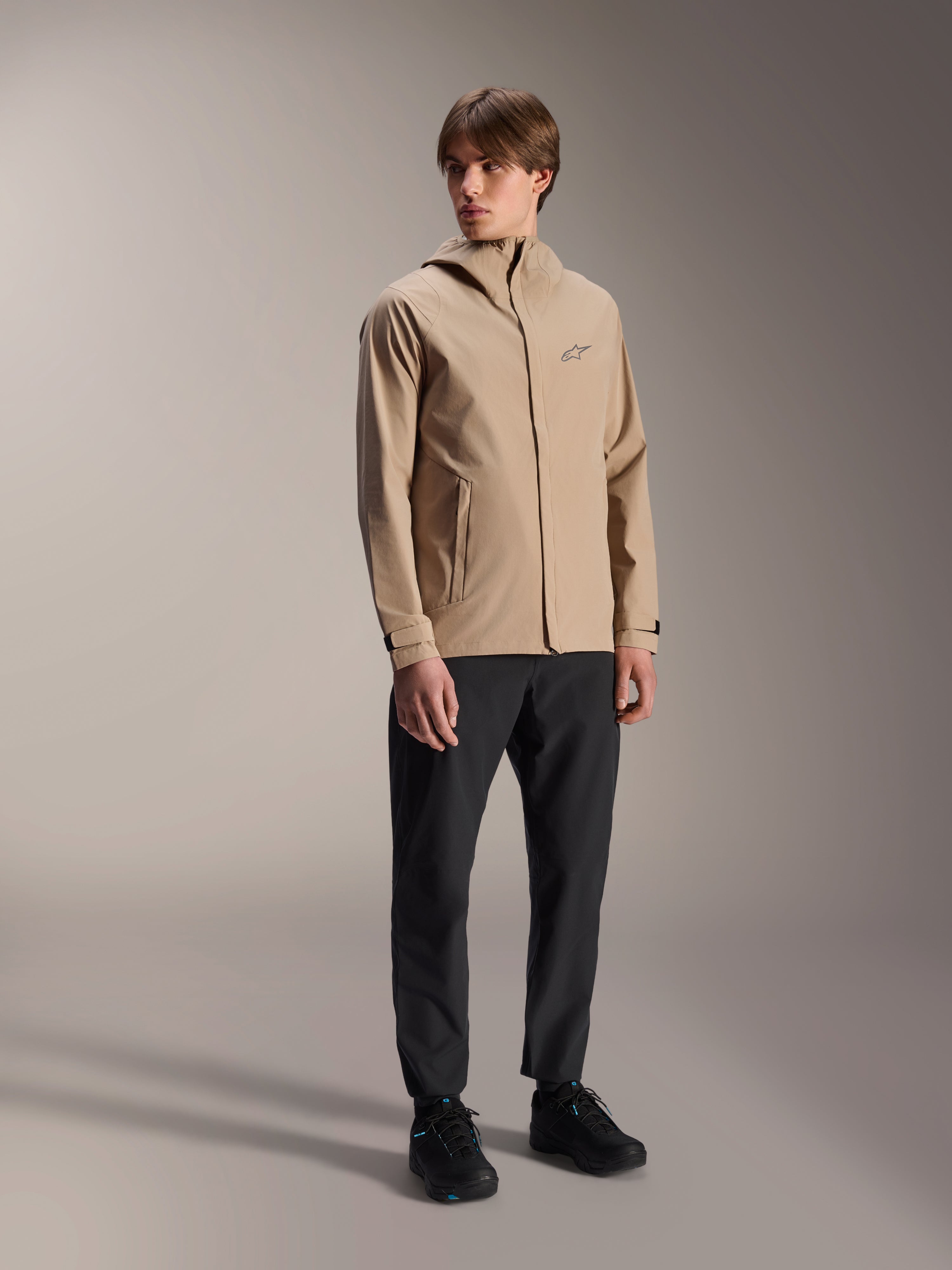 Modello uomo che indossa la giacca antipioggia Alpinestars A-Dura in Light Brown, abbinata a pantaloni e scarpe neri, giacca antipioggia minimalista per mountain bike outdoor con design con cappuccio e un piccolo logo sul petto.