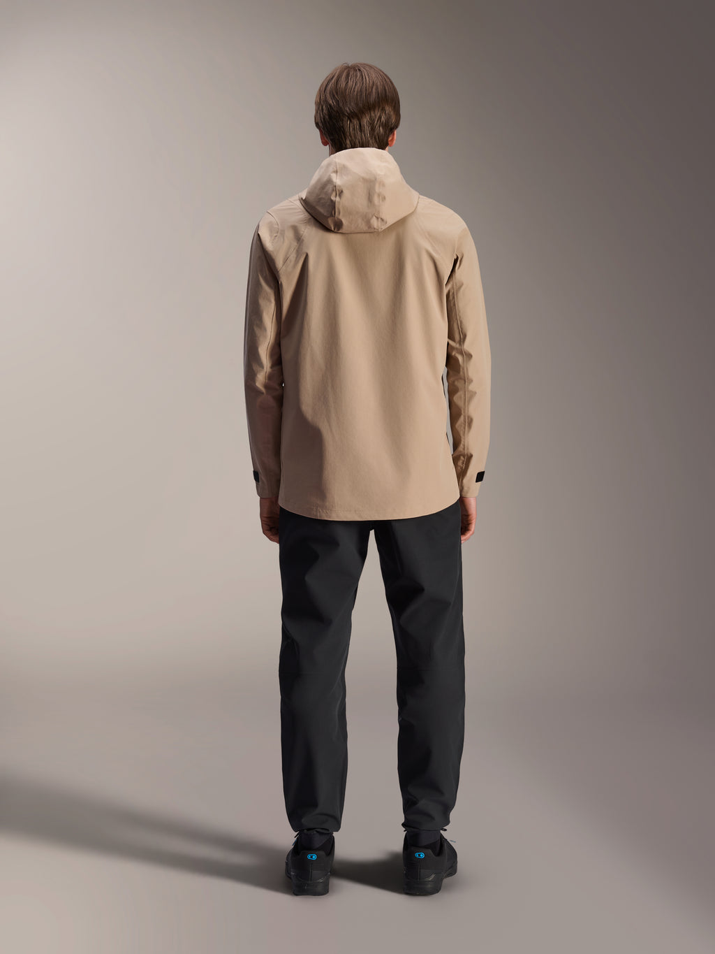 Giacca antipioggia Alpinestars A-Dura, marrone chiaro, vista posteriore con cappuccio e polsini regolabili, indossata con pantaloni neri, abbigliamento versatile da MTB per ogni condizione atmosferica
