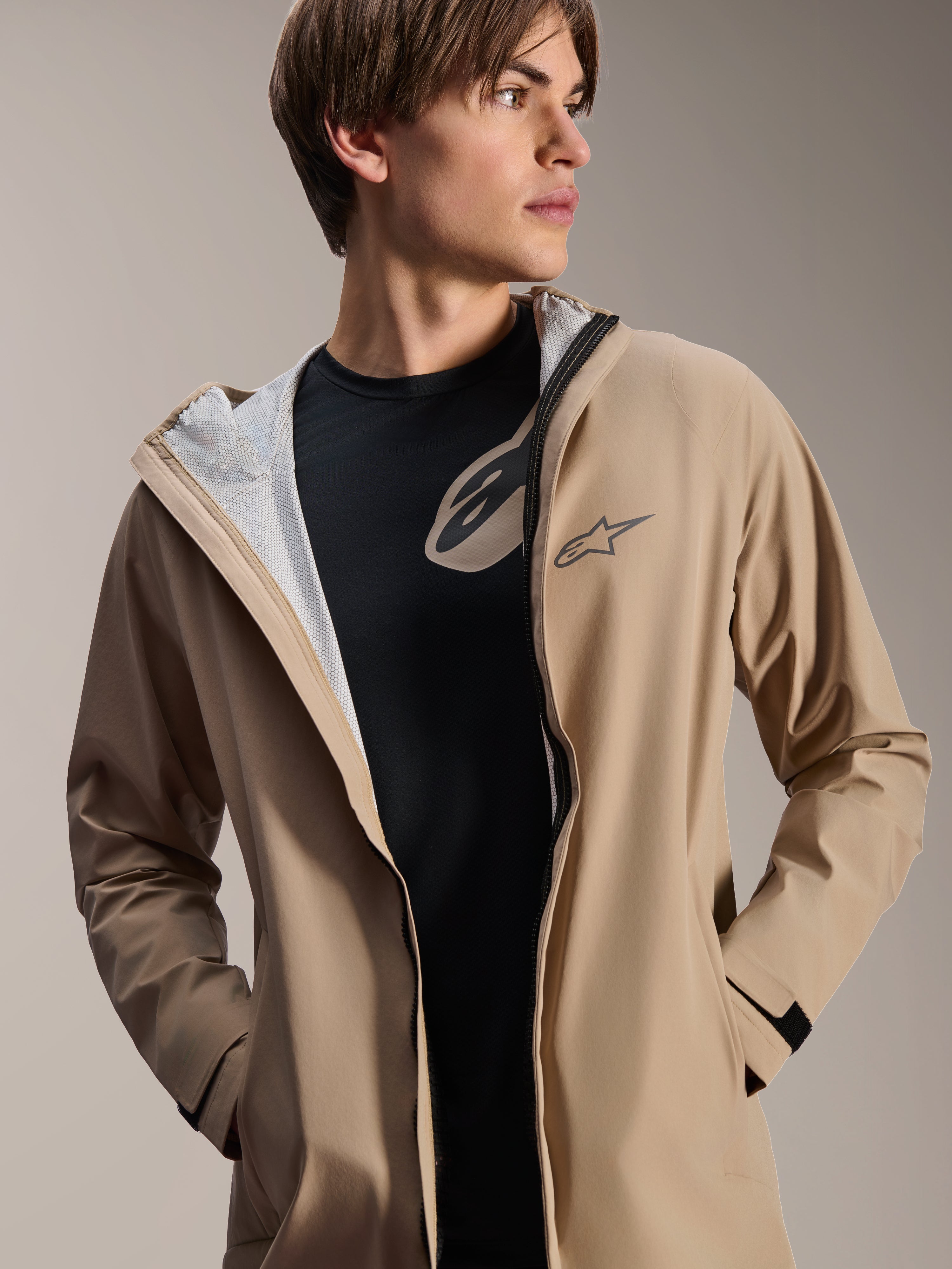 Modello uomo che indossa la giacca antipioggia Alpinestars A-Dura, marrone chiaro, aperta sopra una t-shirt nera, con fodera in rete bianca visibile, giacca da ciclismo versatile e resistente alle intemperie