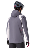 Alpinestars Giacca Antipioggia A-Dura, giacca da mountain bike, grigio titanio scuro, vista posteriore con pannelli su spalle e braccia grigio chiaro, dettaglio logo rosso sul lato, indossata con casco MTB grigio e guanti neri e rossi, abbigliamento tecnico da trail riding