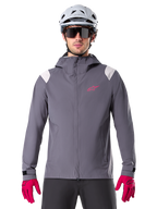 Alpinestars A-Dura Rain Jacket, giacca antipioggia da mountain bike, grigio Dark Titanium, design con cappuccio con logo Alpinestars rosa e inserti grigio chiaro sulle spalle, indossata con casco da MTB bianco e guanti rosa acceso, abbigliamento impermeabile per ciclismo fuoristrada