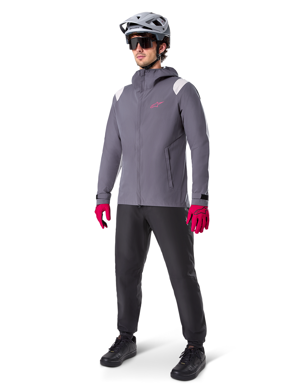 Modello che indossa la giacca antipioggia Alpinestars A-Dura in Dark Titanium, giacca grigia con cappuccio, logo rosa sul petto e inserti grigio chiaro sulle spalle, abbinata a un casco da MTB grigio, occhiali da sole scuri, guanti rosa vivace e pantaloni neri per ciclismo fuoristrada.