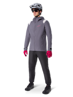 Modello che indossa la giacca antipioggia Alpinestars A-Dura in Dark Titanium, giacca grigia con cappuccio, logo rosa sul petto e inserti grigio chiaro sulle spalle, abbinata a un casco da MTB grigio, occhiali da sole scuri, guanti rosa vivace e pantaloni neri per ciclismo fuoristrada.