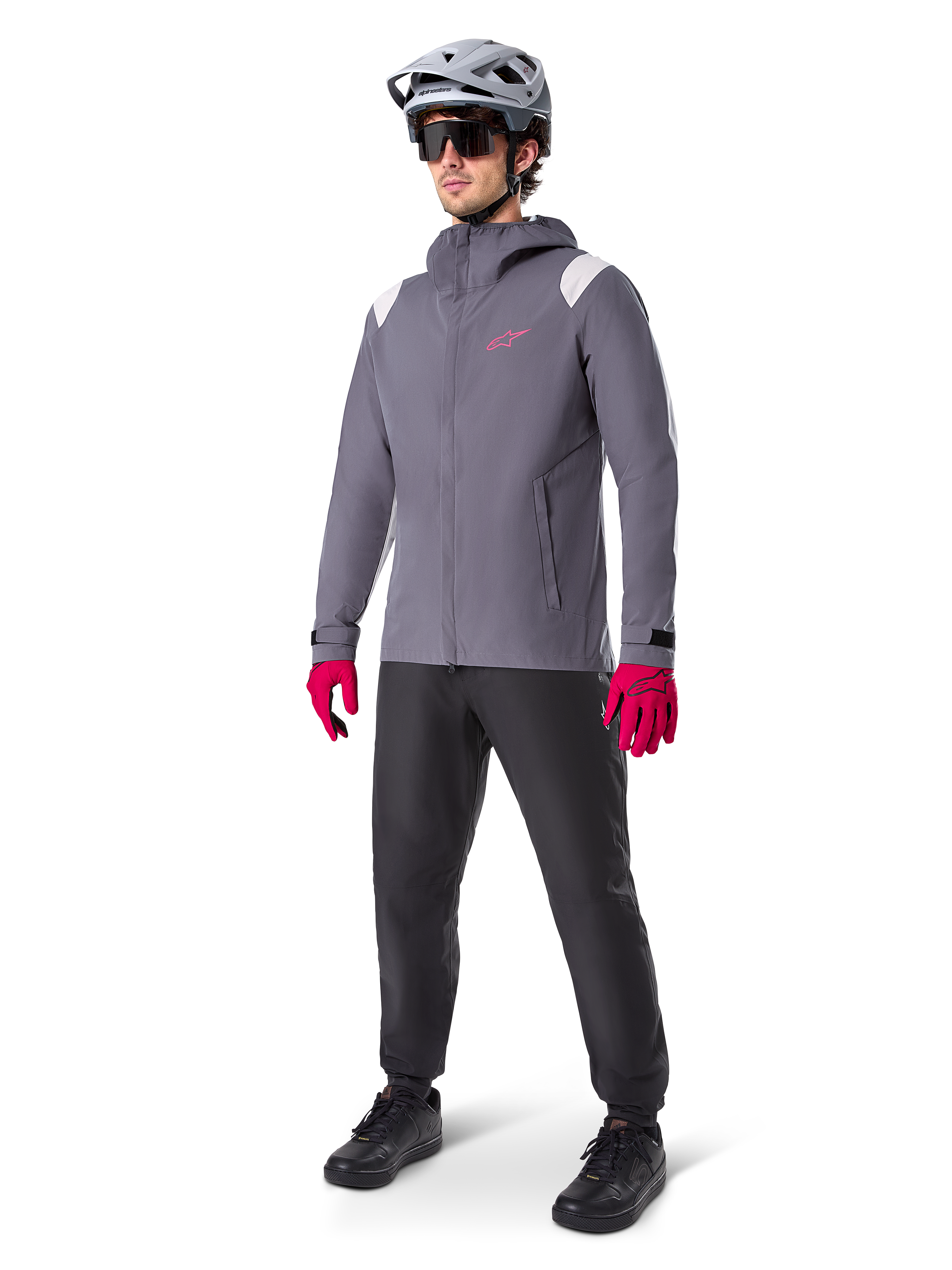 Modello che indossa la giacca antipioggia Alpinestars A-Dura in Dark Titanium, giacca grigia con cappuccio, logo rosa sul petto e inserti grigio chiaro sulle spalle, abbinata a un casco da MTB grigio, occhiali da sole scuri, guanti rosa vivace e pantaloni neri per ciclismo fuoristrada.