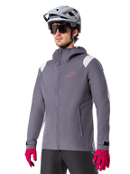 Alpinestars A-Dura Rain Jacket, giacca antipioggia da mountain bike, grigio titanio scuro con logo rosa e accenti bianchi sulle spalle, design con cappuccio e tasche anteriori, indossata con guanti rosa e casco da ciclismo grigio chiaro