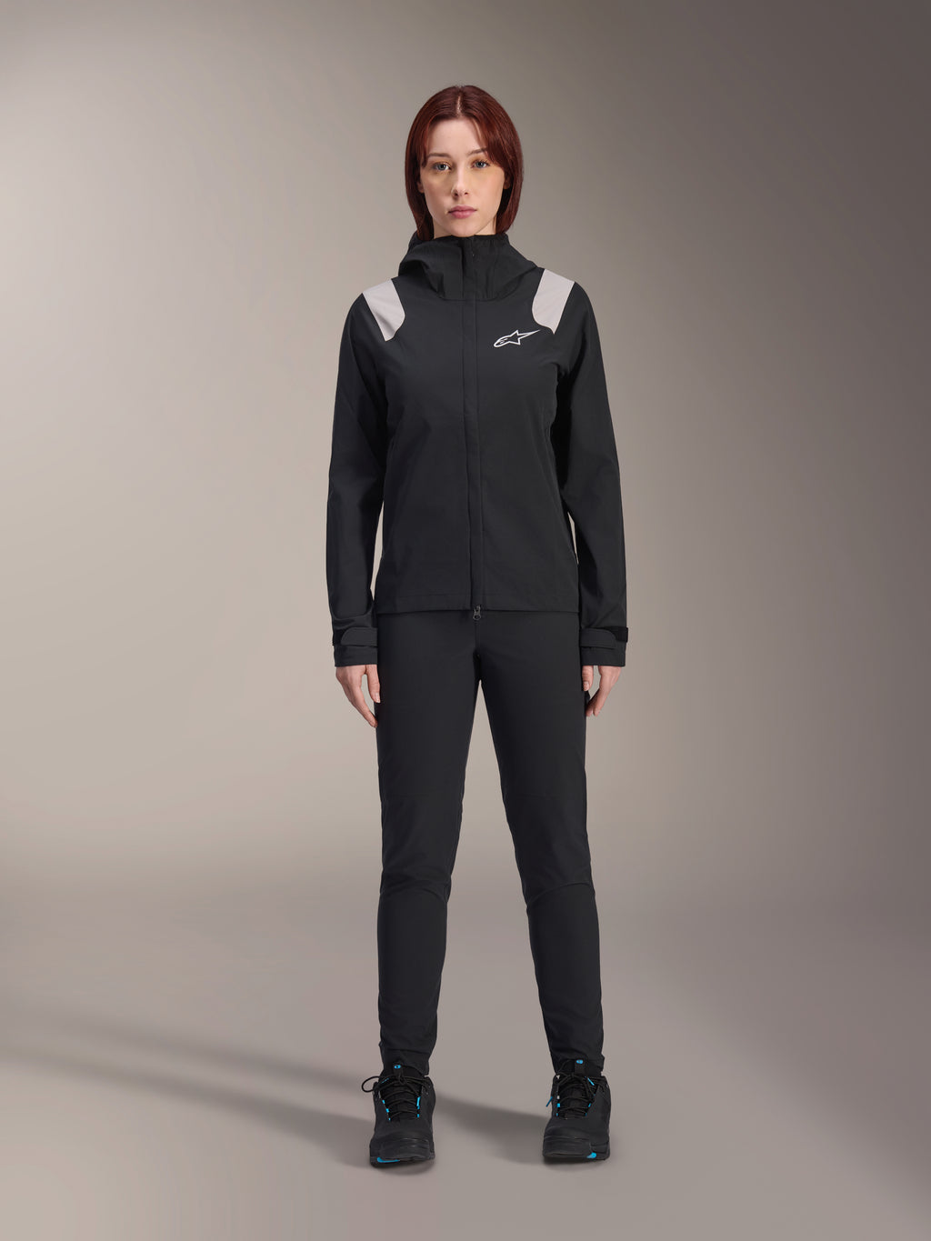 Alpinestars Stella A-Dura Rain Jacket, giacca antipioggia MTB da donna, nera con accenti grigio chiaro sulle spalle e logo bianco, indossata con pantaloni e scarpe neri abbinati, dotata di cappuccio e polsini regolabili, progettata per il trail riding in mountain bike