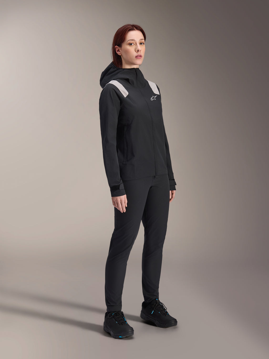 Donna che indossa la giacca antipioggia Alpinestars Stella A-Dura in nero, guscio MTB impermeabile a 2,5 strati con dettagli grigio chiaro sulle spalle, pantaloni neri e scarpe da ciclismo abbinati, ritratto in studio per abbigliamento tecnico da ciclismo