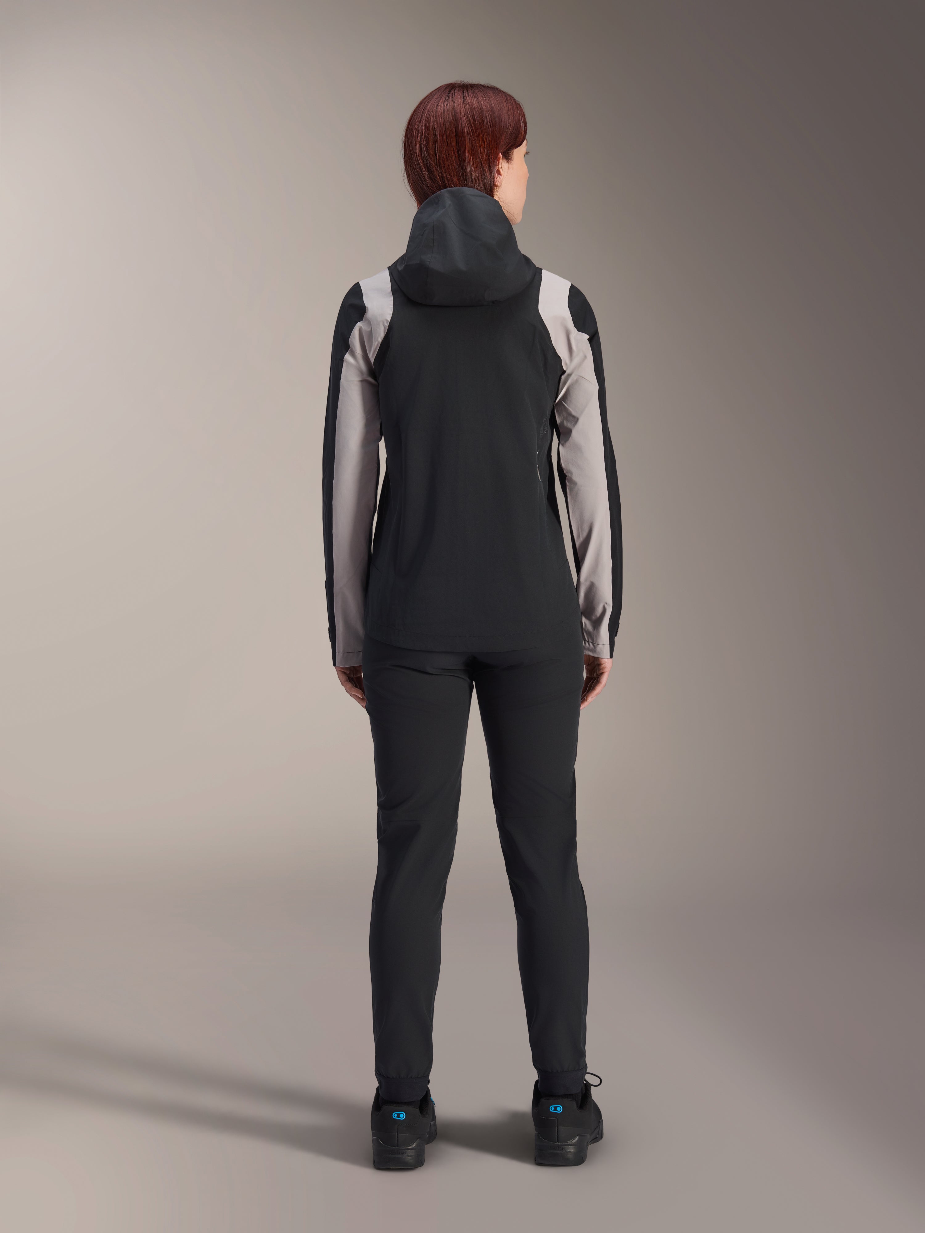 Alpinestars Stella A-Dura Rain Jacket, ciclista vista da dietro, nera con inserti grigio chiaro su maniche e spalle, cappuccio sollevato, indossata con pantaloni neri e scarpe da ciclismo, abbigliamento tecnico antipioggia per mountain bike