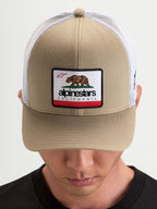 Alpinestars Cappello Cali 2.0, cappello trucker sportswear, kaki e bianco, con pannello frontale in twill strutturato, patch con orso della California e retro in rete traspirante indossato da un modello