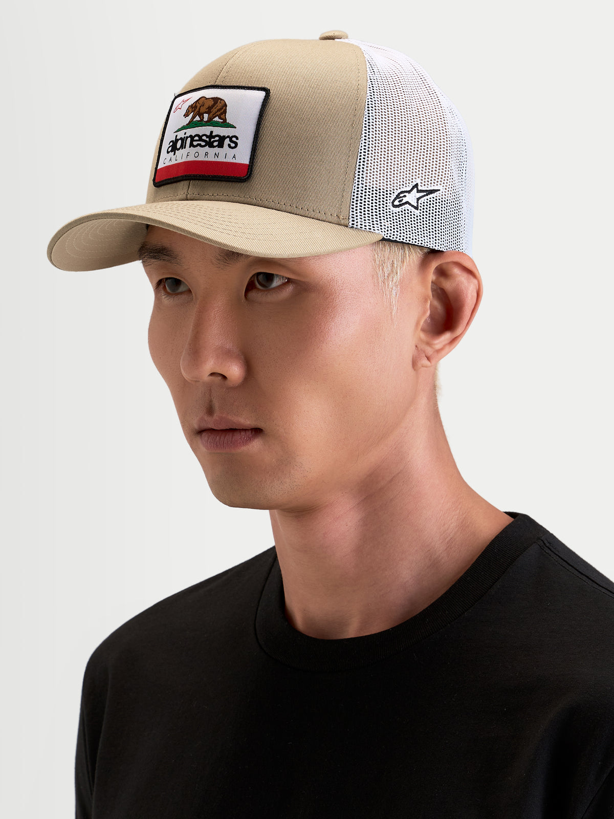 Alpinestars Cappello Cali 2.0, cappello sportswear trucker in khaki e bianco, indossato da un modello, con visiera curva, corona strutturata in twill, retro in rete traspirante e patch California sul davanti