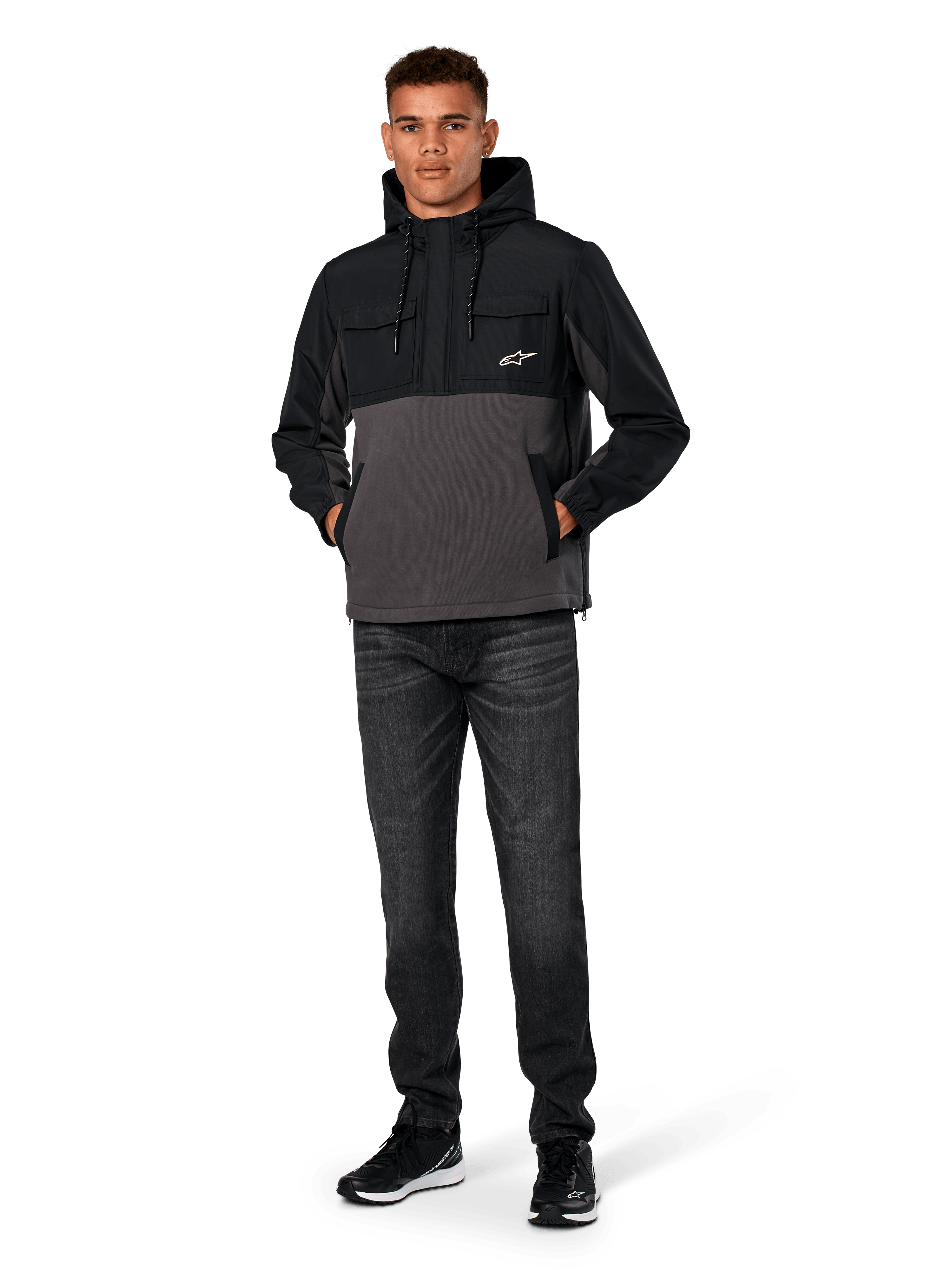 Giacca Hybrid Alpinestars Juncture, nero e gunmetal, felpa anorak con cappuccio in materiali misti, corpo in tela e pile con sovrapposizioni in ripstop, logo A-star bianco sul petto, cappuccio con coulisse, indossata da un modello con jeans scuri e sneakers nere, streetwear casual ispirato al motorsport