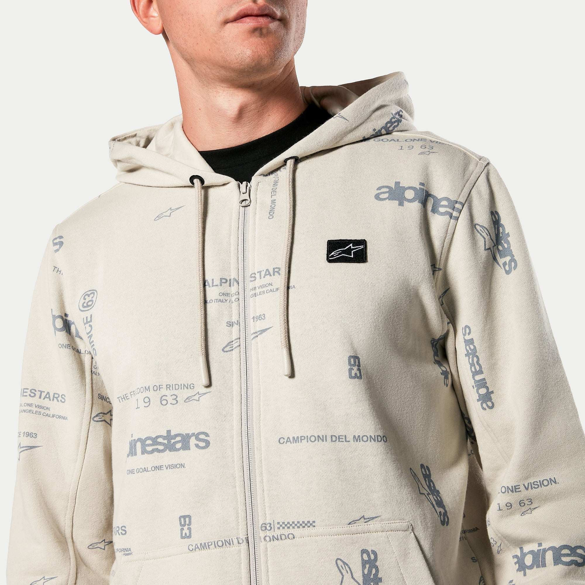 Felpa con cappuccio Alpinestars Plenitude, pile con zip frontale in Off White, stampa grafica integrale con testo 'alpinestars' e '1963', patch logo nera ricamata sul petto, cappuccio con cordino, streetwear casual ispirato al motorsport