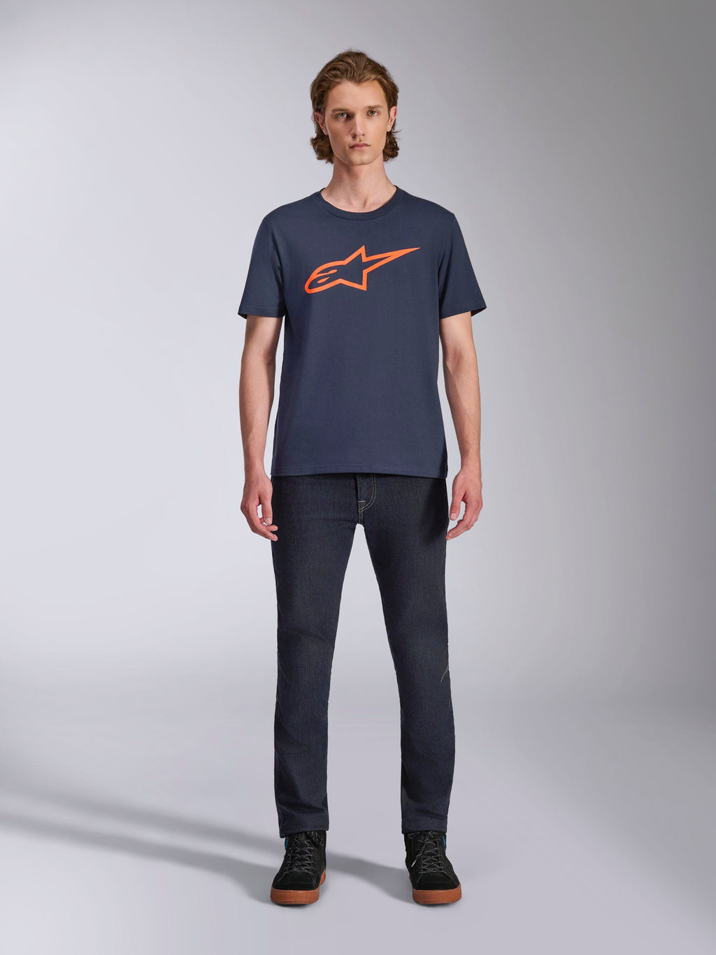 Alpinestars Ageless 2.0 CSF Tee, maglietta a maniche corte, blu navy con logo Alpinestars arancione, indossata da un modello con jeans scuri e sneakers, streetwear casual da motorsport