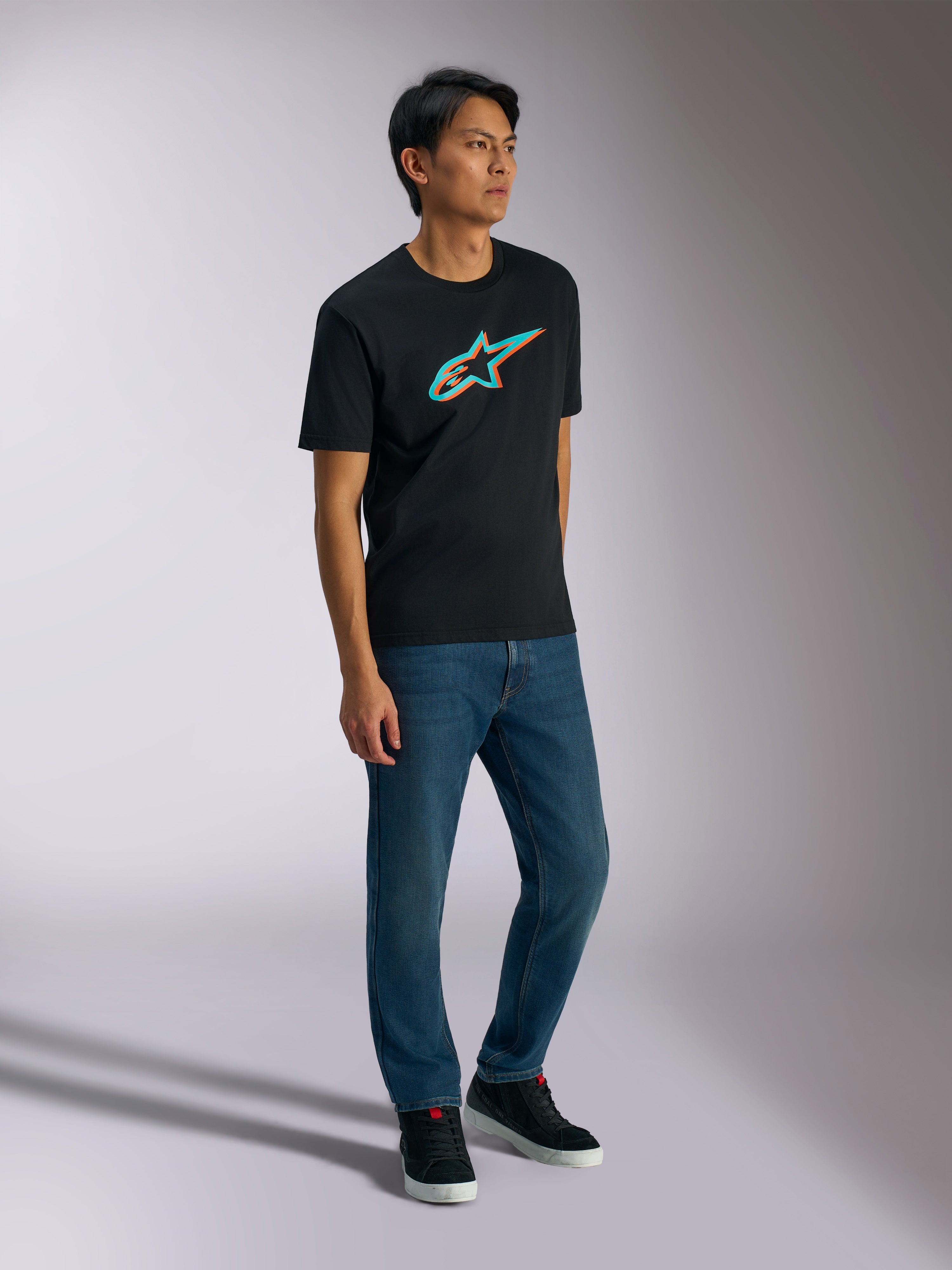 Alpinestars Ageless Shadow CSF Tee, maglietta a maniche corte, nero con logo in ombra turchese e arancione, indossata da un modello uomo con jeans blu e sneakers nere, streetwear casual ispirato al motorsport