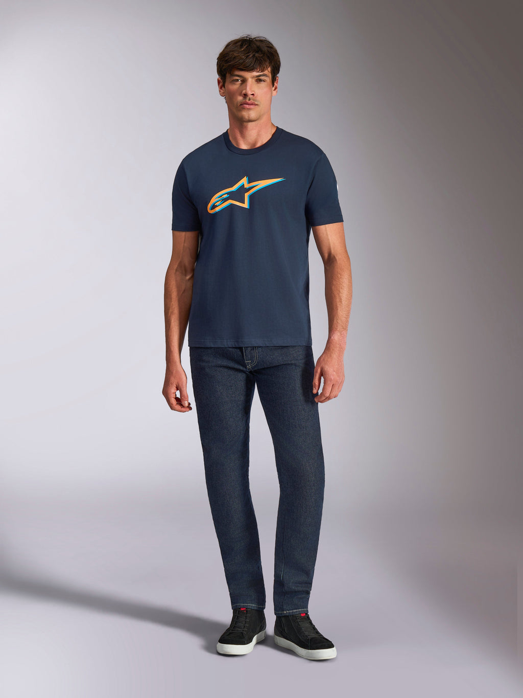 Alpinestars Ageless Shadow CSF Tee, t-shirt a maniche corte, blu navy, con logo Ageless Shadow arancione e azzurro, indossata da un modello con jeans in denim scuro e sneakers nere, streetwear casual ispirato al motorsport