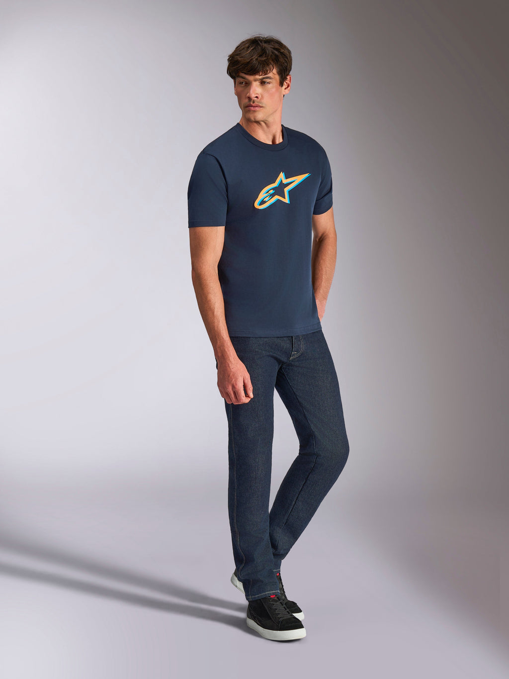 Alpinestars Ageless Shadow CSF Tee, maglietta a maniche corte, blu navy con logo in ombra arancione e verde acqua, indossata da un modello con jeans in denim scuro e sneakers nere, streetwear casual ispirato al motorsport