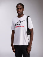 Alpinestars Always 2.0 CSF Tee, T-shirt a maniche corte, bianca con grande logo a stella nero e logo rosso, indossata da un modello con pantaloni neri e borsa a tracolla, streetwear casual ispirato al motorsport