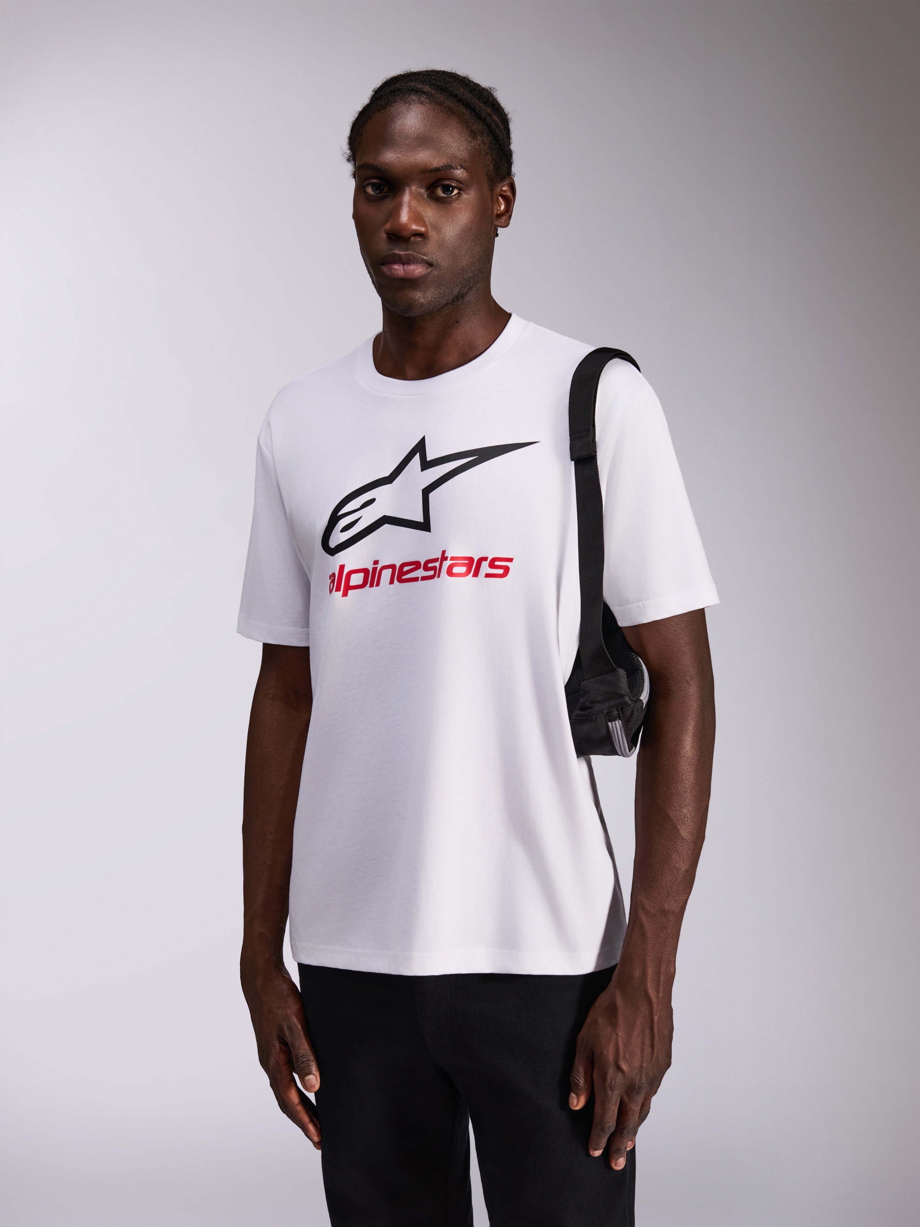 Alpinestars Always 2.0 CSF Tee, T-shirt a maniche corte, bianca con grande logo a stella nero e logo rosso, indossata da un modello con pantaloni neri e borsa a tracolla, streetwear casual ispirato al motorsport