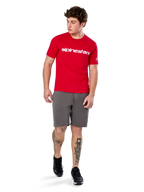 Alpinestars Linear Wordmark 2.0 CSF Tee, t-shirt a maniche corte, rossa con logo wordmark bianco sul petto, indossata da un modello con pantaloncini grigi e sneakers nere, streetwear casual ispirato al motorsport