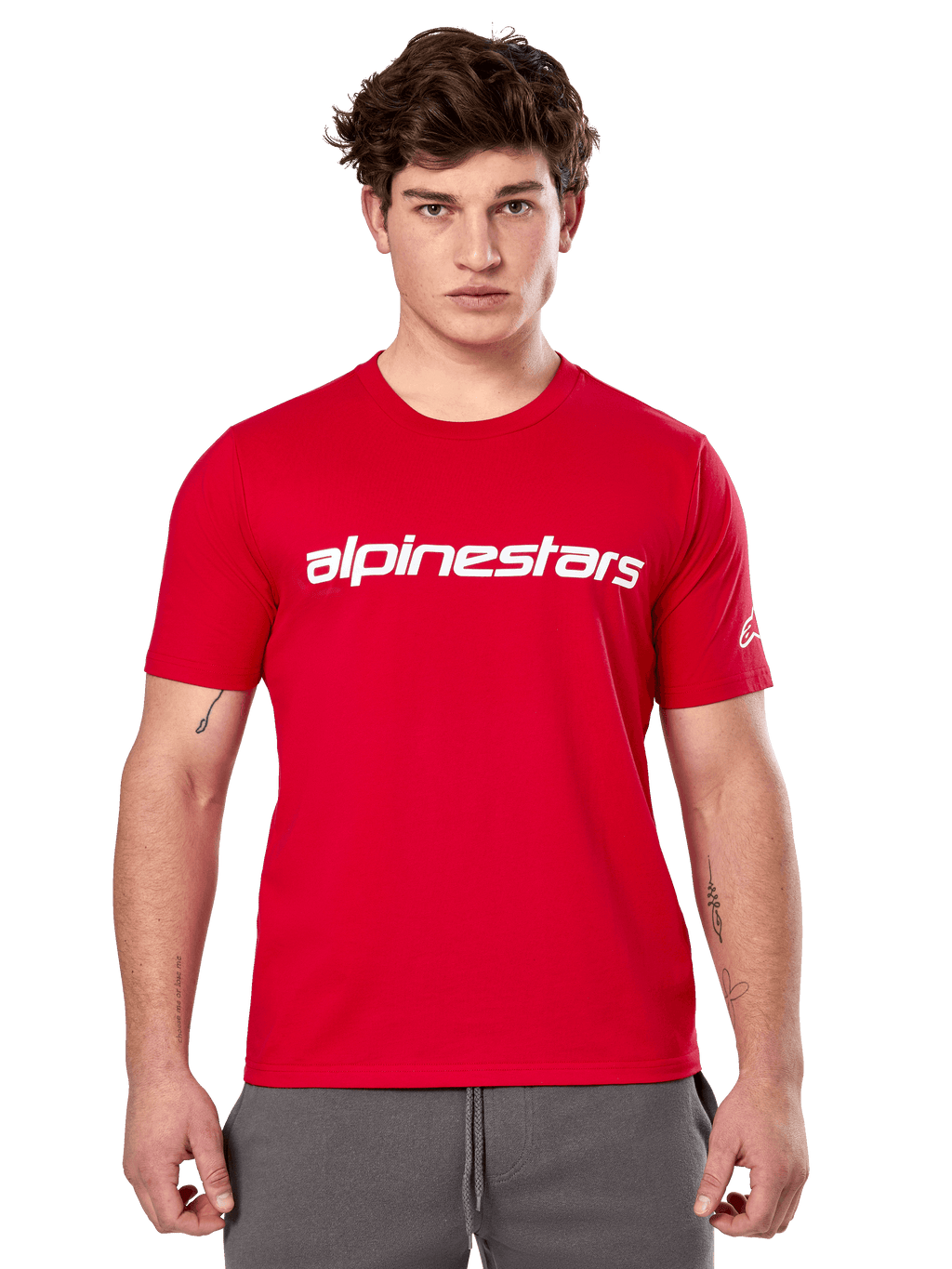 Alpinestars Linear Wordmark 2.0 CSF Tee, maglietta a maniche corte, rossa, logo lineare bianco sul petto e logo Ageless sulla manica, indossata da un modello con pantaloni della tuta grigi, streetwear casual ispirato al motorsport